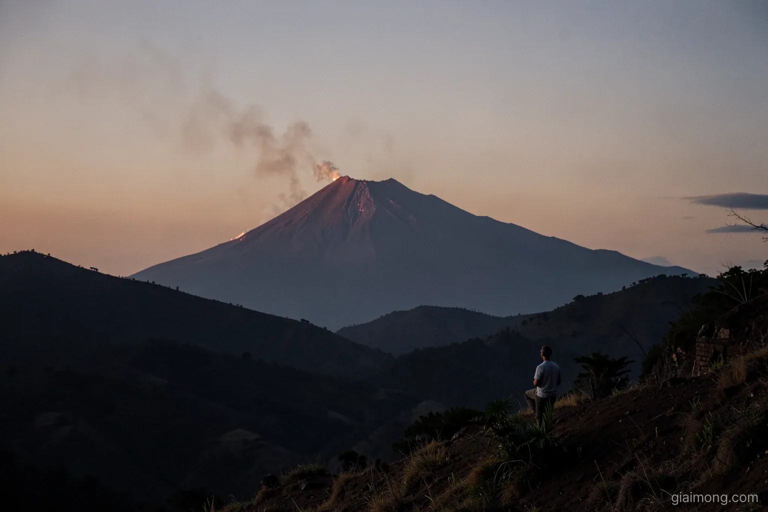 Distant volcanic cone glowing faintly orange at dusk above dark rolling hills, a thin plume of smoke drifting, a lone figure watching from a hillside